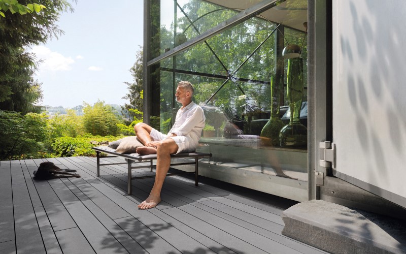 man wearing white sitting behind a glass window grey facade and green trees with light blue sky on a left