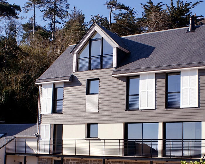 big grey with white sidings house dark grey roof and dark trees behind it
