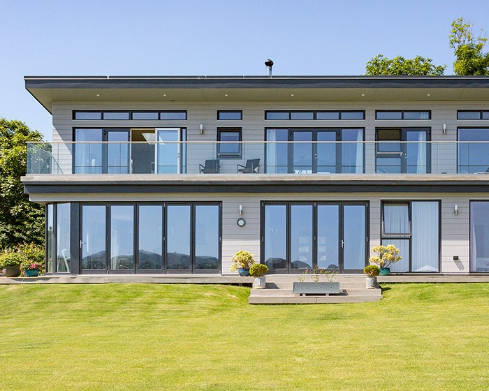 green grass white sidings house behind with many big windows above it the sky is light blue