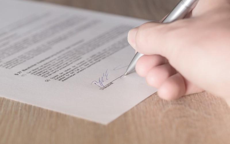 white paper being signed with blue ink on a right a hand holding silver pen 