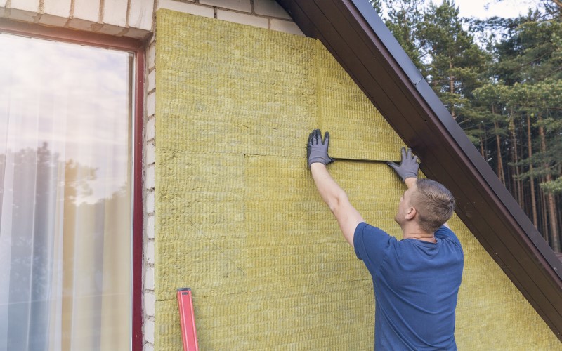 yellow exterior siding being applied by an installer dressed blue on the left a window on the right green forest trees