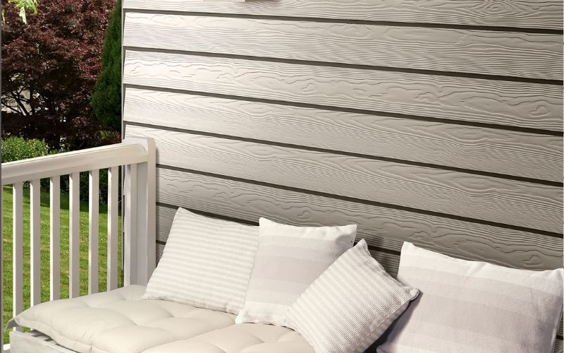 white bed with pillows behind it white siding wall on the left green trees and grass