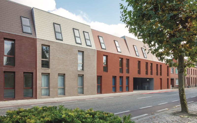 a horizon of houses from the left brown, light grey, orange with a light blue sky on the right green trees and grey asphalt road