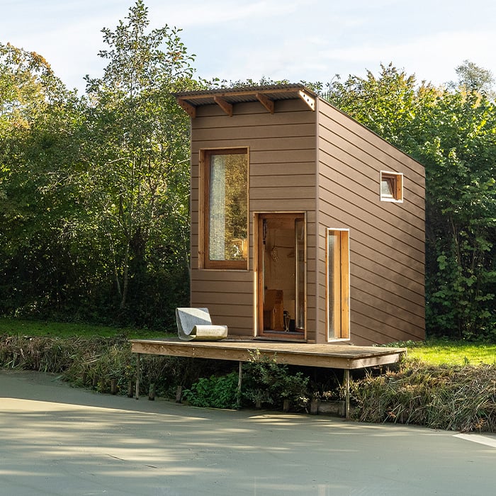 Helle, braune Fassadenverkleidung einer kleinen Hütte direkt am See. Die natürliche Farbgebung fügt sich harmonisch in die Umgebung ein und unterstreicht den ruhigen, naturnahen Charakter des Ortes